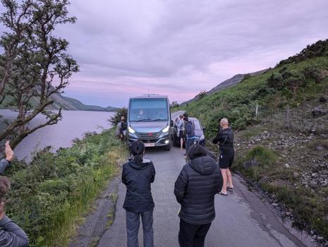 Atmos' coach navigating a tight gap on a single lane road with a broken down car on one side and a lake on the other