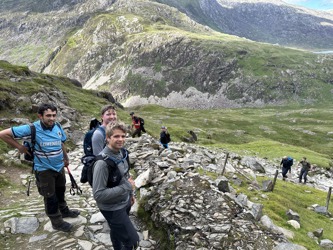 A line of people climbing one of the three peaks in the background. Three Atmos colleagues are in the foreground smiling at the camera